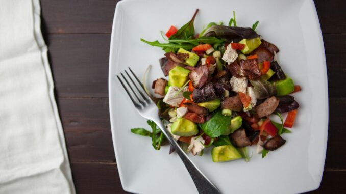 A fresh bacon, avocado, and mixed greens salad served on a square plate with a fork.